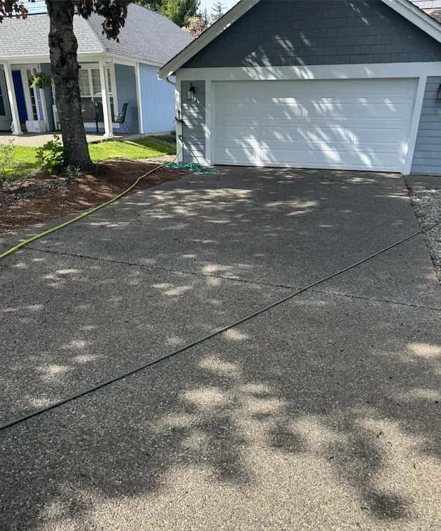 Dirty exposed-aggregate driveway in dappled shade leading to a white two-car garage, hoses laid out before washing.