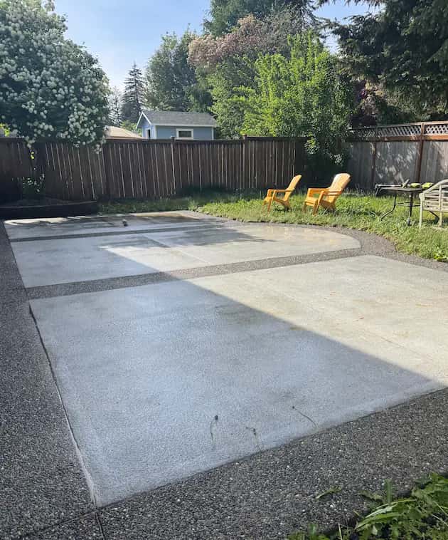 Clean backyard concrete patio with exposed-aggregate borders after pressure washing, yellow Adirondack chairs beyond.