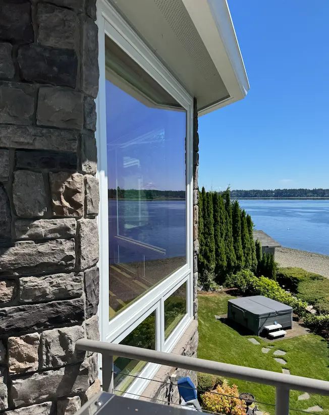 Streak-free corner window on a stone-veneer home reflecting blue sky, overlooking a lawn, hot tub and waterfront.