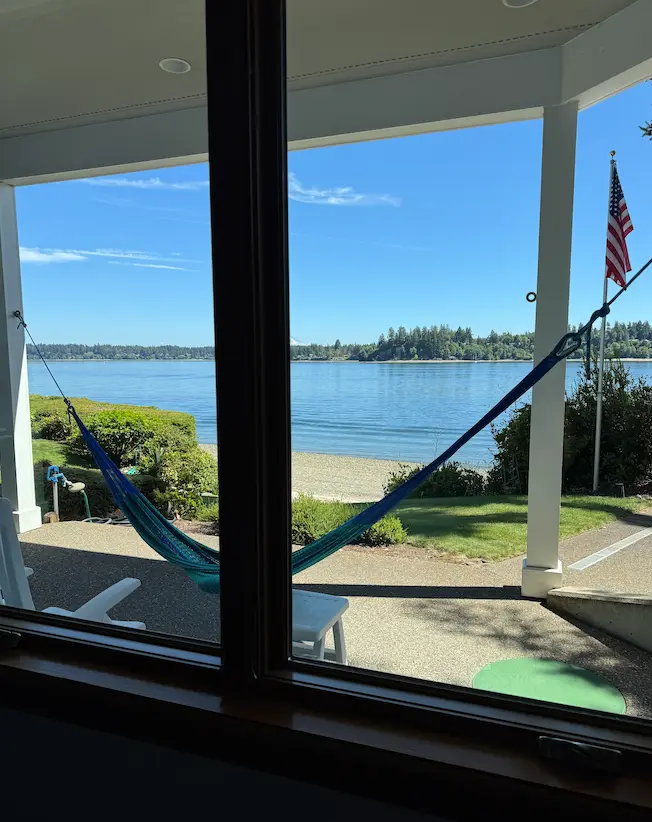 Streak-free interior window looking out on a porch hammock, beach, and calm blue inlet with a treed shoreline.
