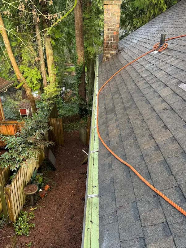 Roof-level view of an emptied gutter along wet asphalt shingles, orange safety rope and brick chimney.