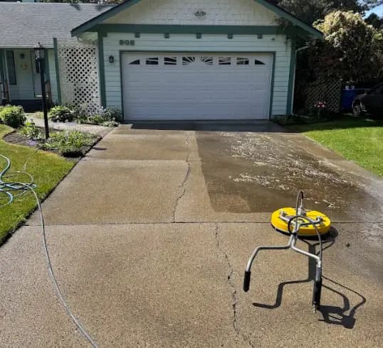 Concrete driveway partly pressure washed, clean strip beside dirty concrete, yellow surface cleaner resting on it.