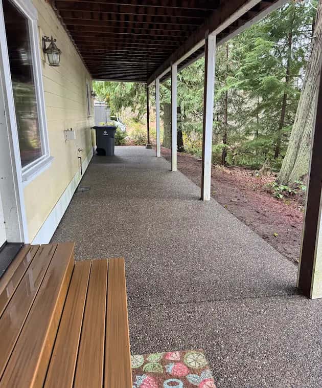 Clean exposed-aggregate patio under a covered deck beside a yellow house, wood bench and evergreens, after pressure washing.