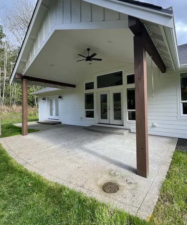 Covered exposed-aggregate patio at a white farmhouse-style home with timber posts, showing stains near the drain.