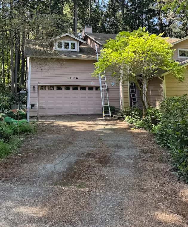 Driveway covered in fir needles and dirt in front of a pink two-story home with ladders against it, before cleaning.