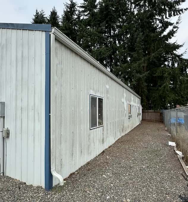 Long white metal-sided building with dirt and streaking along the walls, beside a gravel lot, before washing.