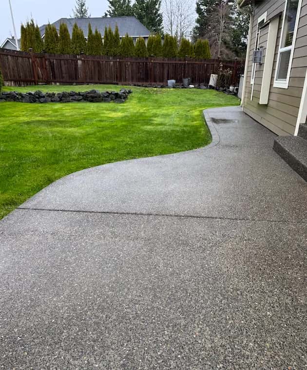 Curved exposed-aggregate concrete patio beside a green lawn and wood fence, freshly cleaned and still damp.