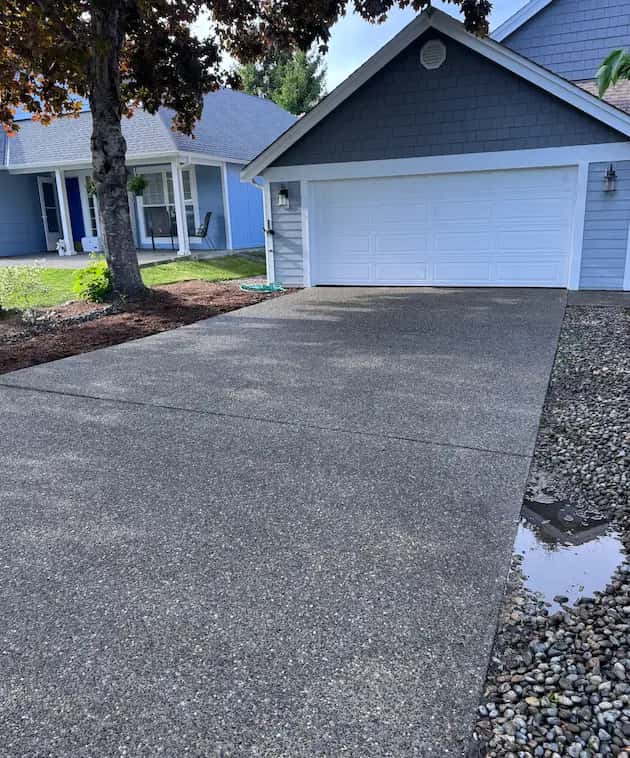 Clean exposed-aggregate concrete driveway leading to a white garage door on a blue house after pressure washing.