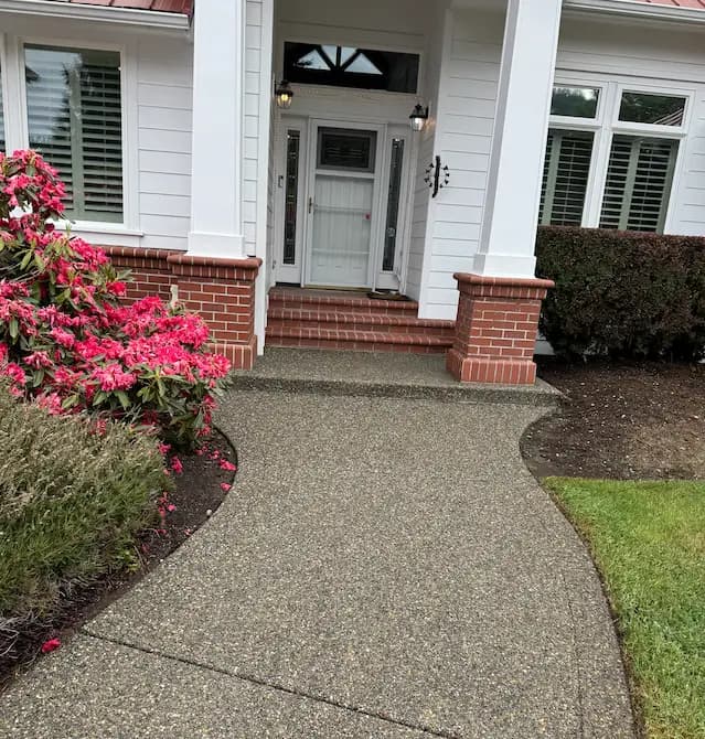Clean exposed-aggregate walkway leading to a white home's brick-trimmed front porch, flanked by pink rhododendrons.
