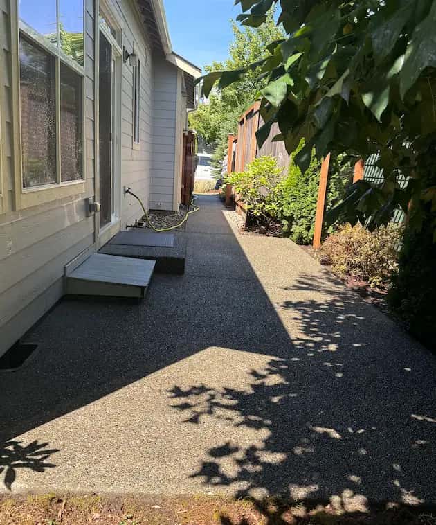Clean exposed-aggregate concrete walkway along the side of a house, bordered by a cedar fence and shrubs.