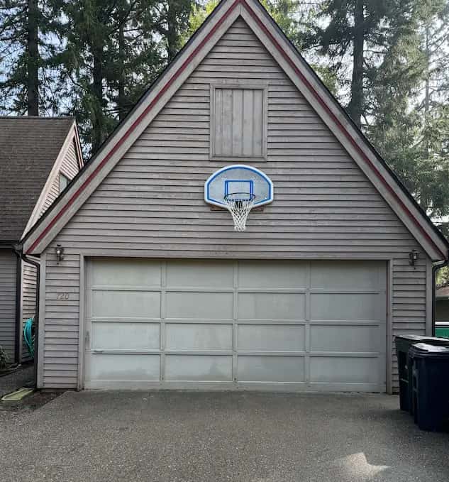 Gabled detached garage with gray lap siding and basketball hoop after washing, clean driveway, tall firs behind.