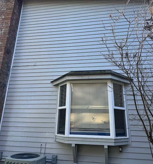 Clean light gray lap siding and white bay window beside a brick chimney after house washing.