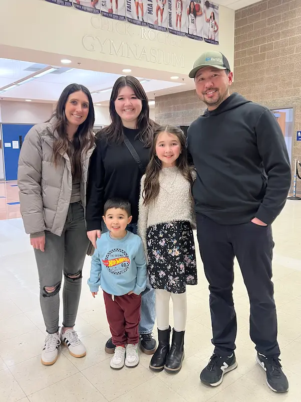 Owner family of five, two adults and three children, smiling together in a school gymnasium lobby.