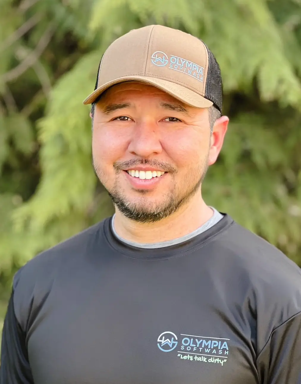Smiling Olympia Softwash owner in a tan company cap and black "Let's talk dirty" logo shirt in front of evergreen trees.