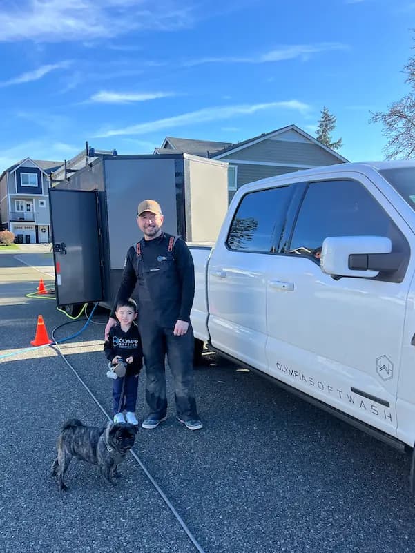Olympia Softwash owner with his young son and a dog beside the white company truck and equipment trailer.