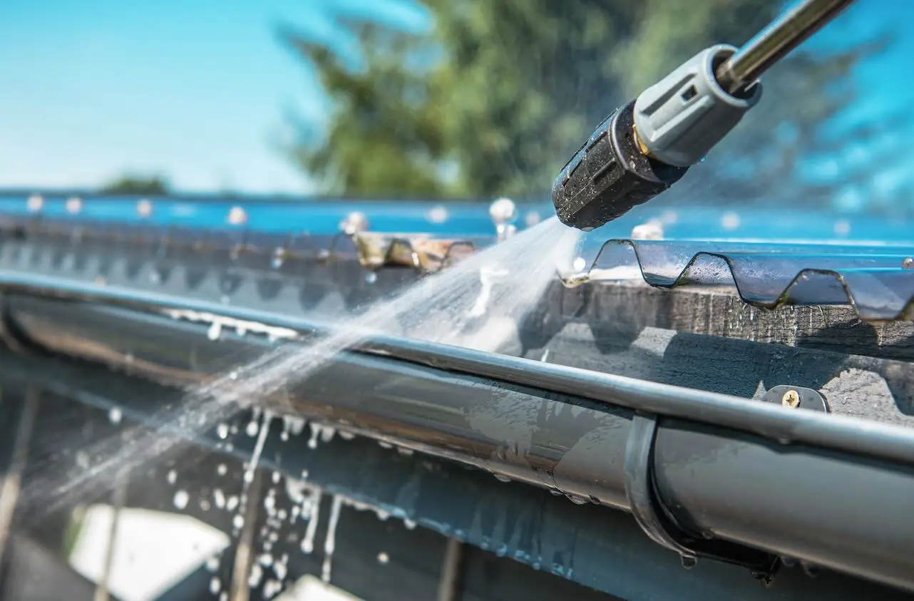 Pressure washer nozzle spraying water along a gray metal rain gutter under a corrugated roof edge.