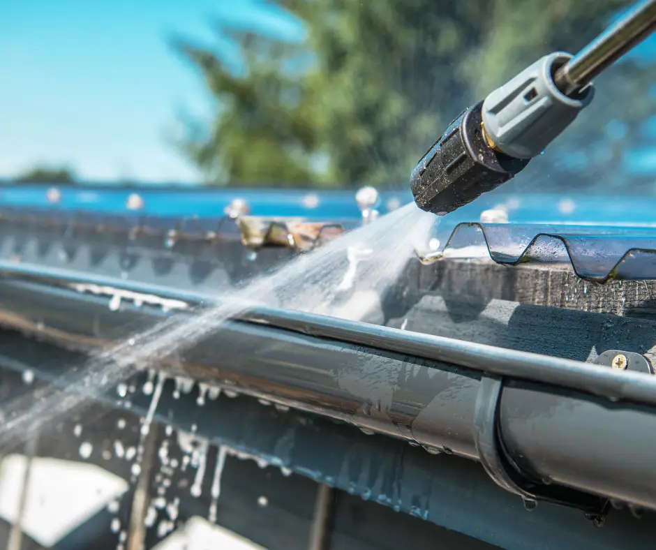 Pressure washer nozzle spraying water along a gray metal rain gutter under a corrugated roof edge on a sunny day.