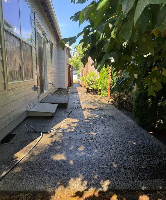 Wet exposed-aggregate side-yard walkway along a house, with hoses laid out during pressure washing.