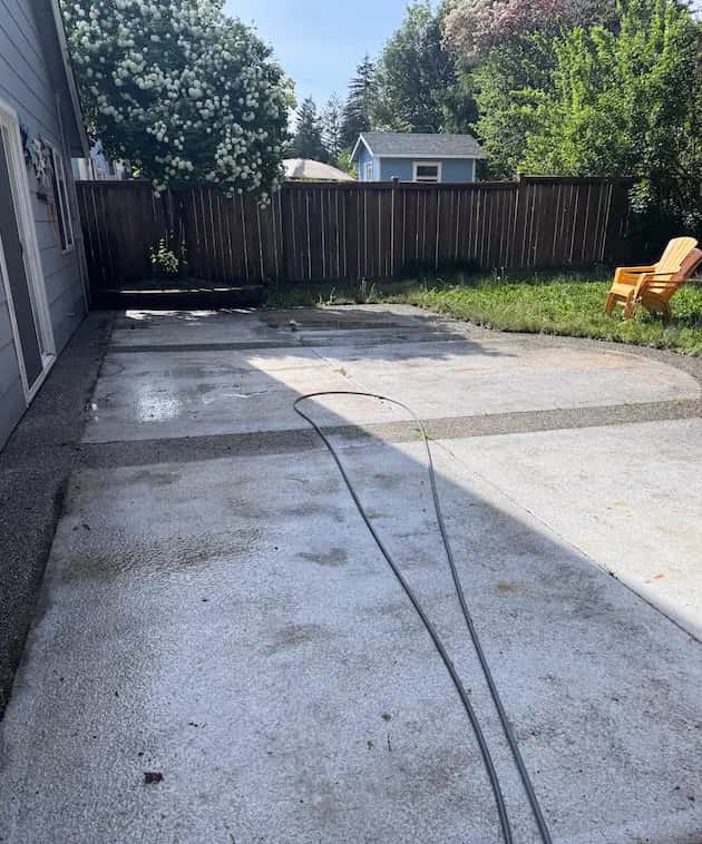 Stained, mildewed concrete backyard patio with a hose on it, wood fence and yellow Adirondack chair, before pressure washing.