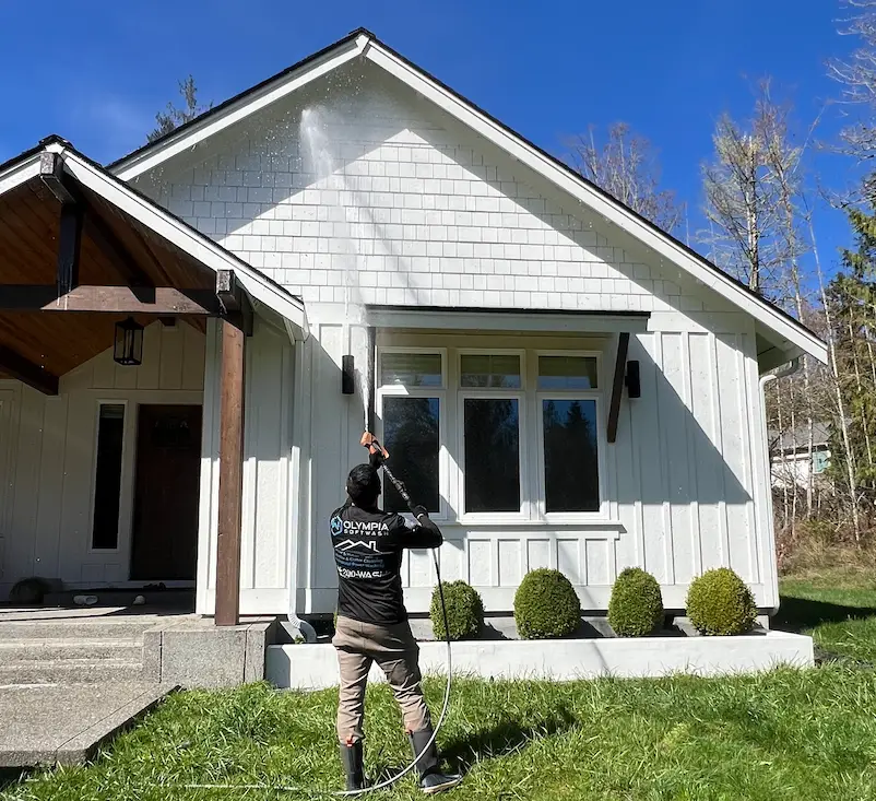 Olympia Softwash technician soft washing the gable of a white board-and-batten house on a sunny day.
