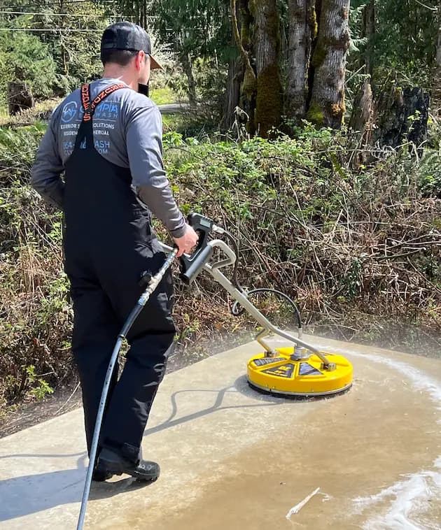 Olympia Softwash technician in bib overalls using a yellow surface cleaner on a concrete pad near woods.