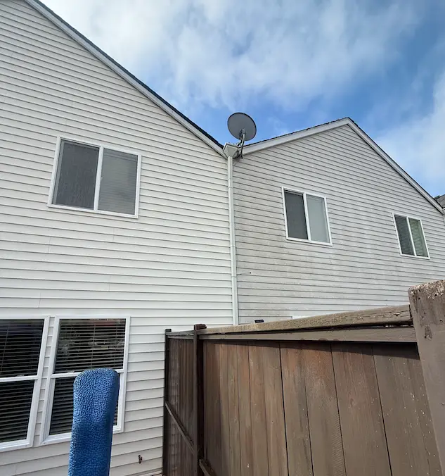 Two-story white vinyl siding mid soft wash: left wall clean, right wall still showing green algae, above a wood fence.