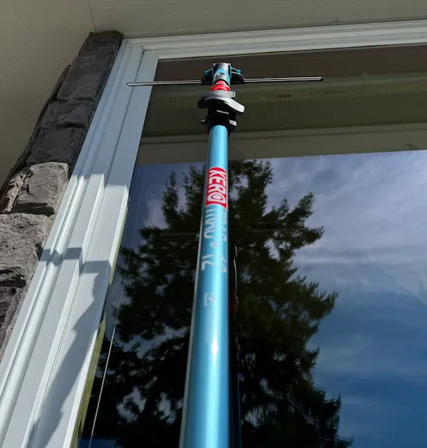 Blue XERO telescoping pole with squeegee against a tall window reflecting an evergreen tree and blue sky.