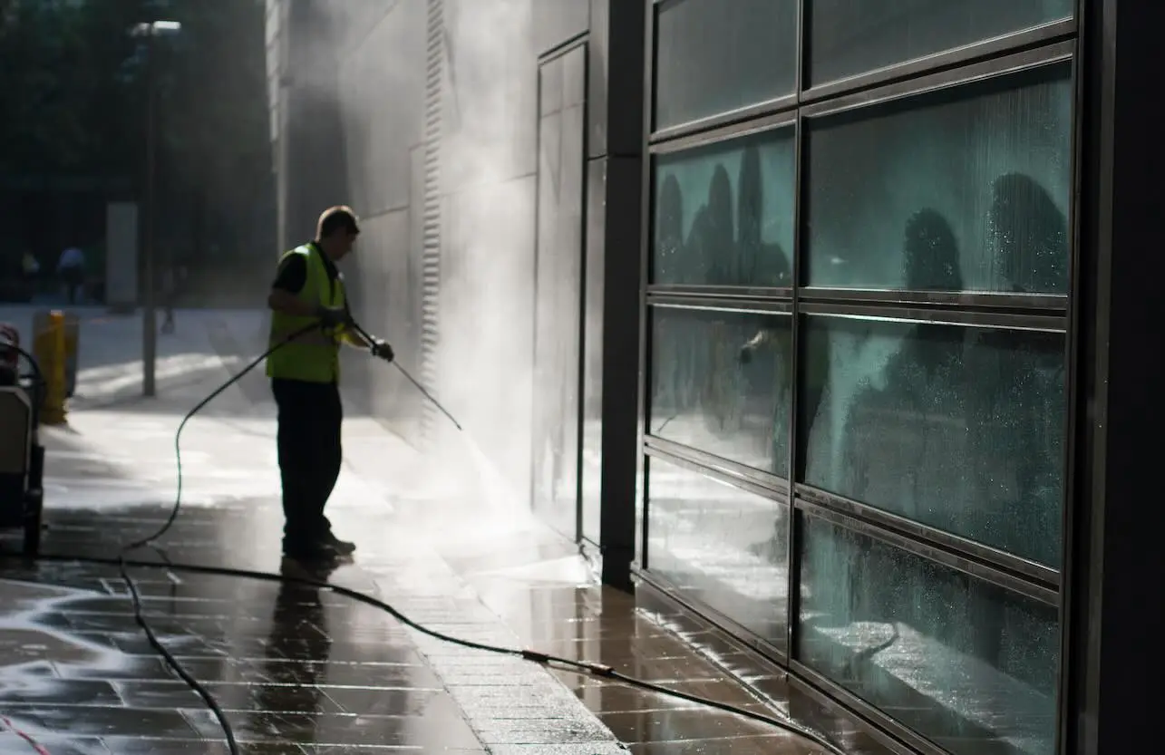 Worker in hi-vis vest pressure washing a commercial building wall and glass facade on a city sidewalk.